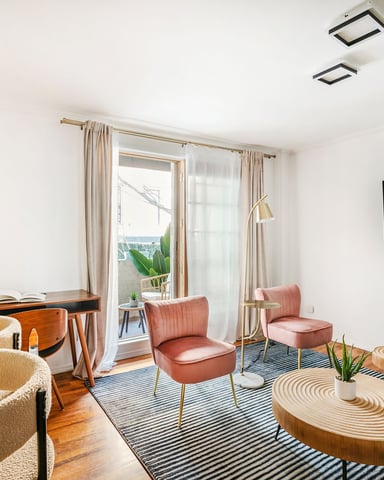 Bright open-plan living room with beige and coral mid-century furniture, wooden desk, wall-mounted TV, and glass doors opening to a balcony.