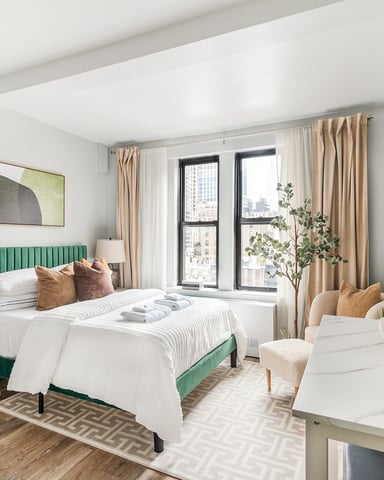 Modern bedroom with green upholstered bed, white bedding, city view windows, and terracotta pottery accents