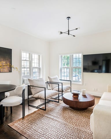 Bright open-plan living room with black oval dining table, cream chairs, wall-mounted TV, and multiple windows with natural light