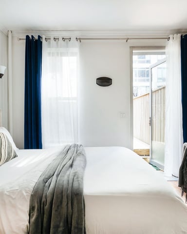 Modern bedroom with queen bed, exposed brick wall, wooden shelving, and glass door opening to balcony