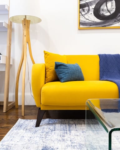 Modern living room with bright yellow sofa, blue and teal pillows, wooden floor lamp, white shelving unit, and framed abstract artwork on white wall.