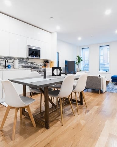Open-plan kitchen and dining area with white cabinetry, dining table with white chairs, blue sofa, and large windows overlooking city skyline.