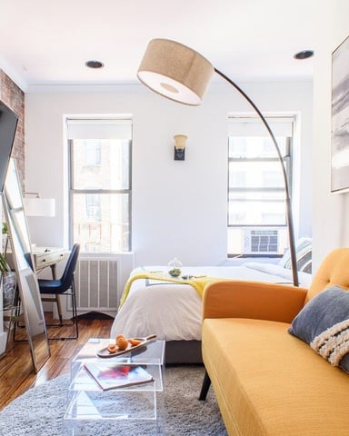 Studio apartment with yellow sofa, bed, wall-mounted TV, arc lamp, two windows, and potted plant on hardwood floor
