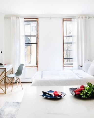 Bright studio apartment with bed, desk area, windows with city views, and kitchen counter with fresh produce