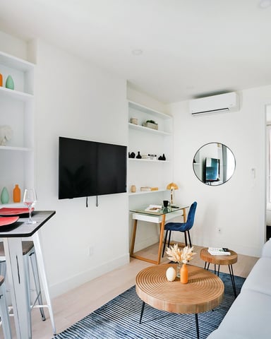 Modern studio apartment with dining table, wall-mounted TV, sofa, and potted plant in bright white interior