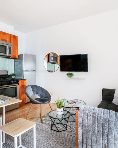 Open-plan kitchen and living room with orange cabinetry, black countertops, wall-mounted TV, and seating area with black sofa and accent chair.