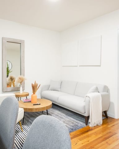 Bright studio apartment with white sofa, round wooden table, gray chairs, hardwood floors, and open doorway to bedroom with blue curtains