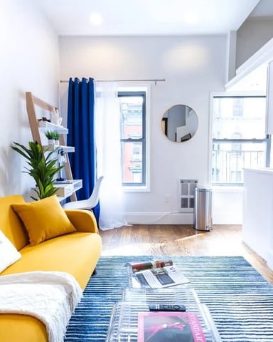 Bright studio apartment with yellow sofa, blue curtains, wall-mounted TV, and city views from balcony door