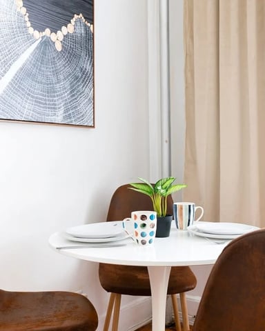 Round white dining table with brown leather chairs, place settings, and potted plant, abstract framed artwork on wall above