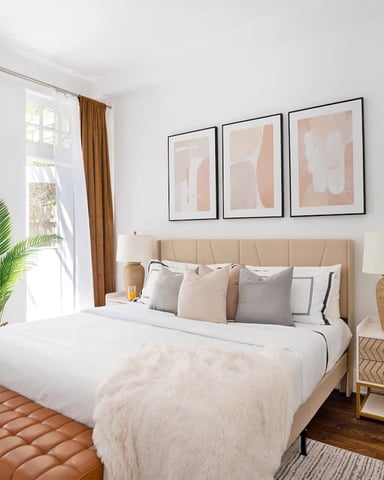 Bright bedroom with beige upholstered bed, three framed abstract artworks, potted plant by window, and wooden nightstand with lamp.