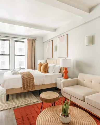 Modern hotel bedroom with queen bed, beige sofa, large windows with city views, and contemporary artwork on walls