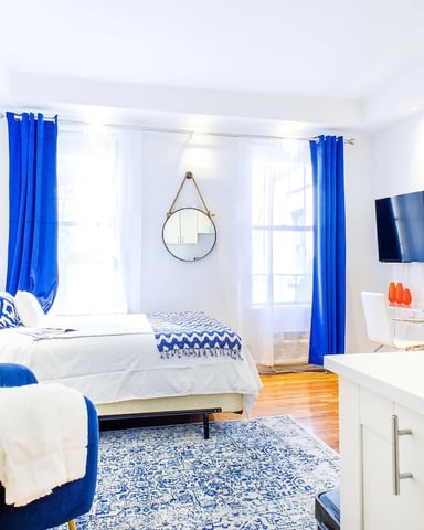 Bright studio apartment with white walls, blue curtains, bed with white bedding, round mirror, TV, potted plant, and hardwood floors.