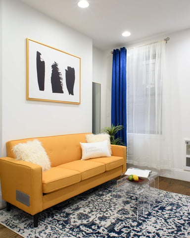 Bright studio apartment with yellow sofa, desk, shelving, abstract wall art, blue curtains, and hardwood floors