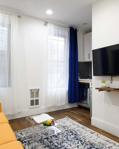Studio apartment living room with yellow sofa, wall-mounted TV, blue curtains, and kitchenette with black brick backsplash