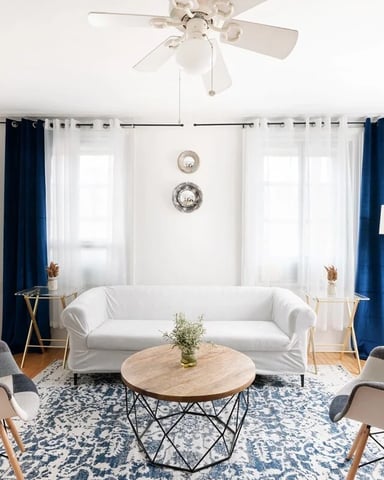 Bright living room with white sofa, two patterned armchairs, round wood table, and blue curtains framing multiple windows