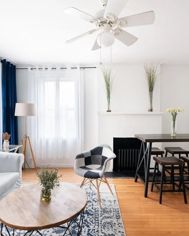 Bright open-plan living room with white sofa, round marble table, black dining set, hardwood floors, and wall-mounted TV.