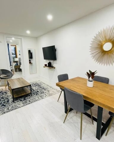 Open-plan living and dining area with wooden table, gray chairs, wall-mounted TV, and gold sunburst mirror