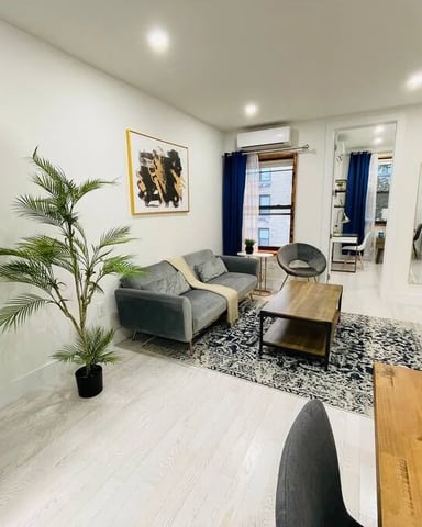 Open-plan studio with stacked washer-dryer, gray seating area, wooden table, potted plant, and wall-mounted TV