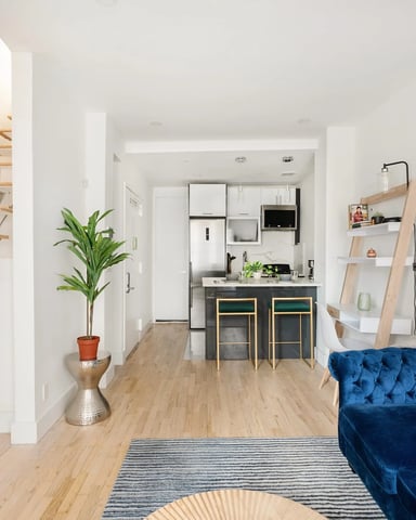 Open-plan living space with blue velvet sofa, wooden floating shelves, kitchen with black island, and wood staircase.