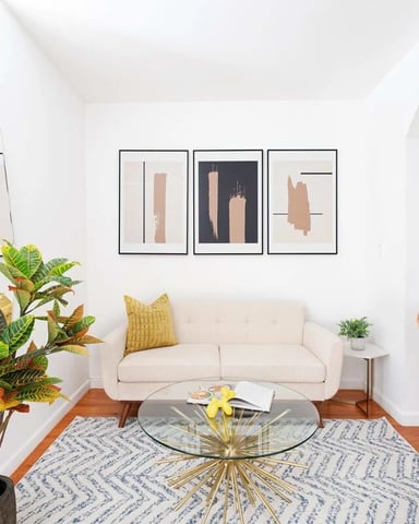 Bright living room with cream sofa, wooden chairs, potted plants, glass coffee table, and abstract wall art