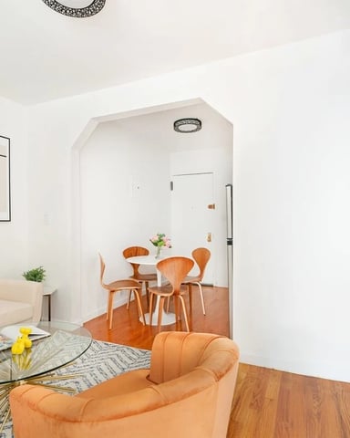 Modern living room with orange chair, glass coffee table, wall-mounted TV, and open-plan dining area with wooden chairs visible through archway