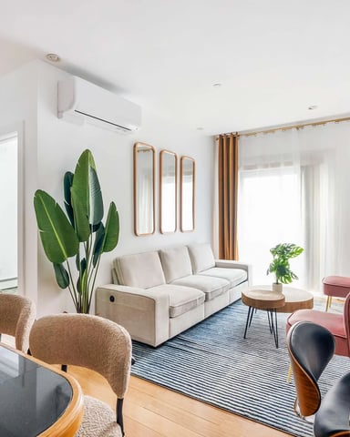Modern living room with beige sofa, pink armchairs, wooden dining table, potted plant, and wall-mounted TV with gold curtains