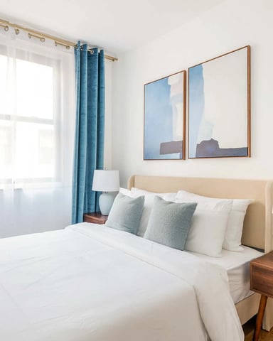 Bright bedroom with beige upholstered bed, white bedding, blue curtains, wooden nightstand, and framed landscape artwork above headboard.
