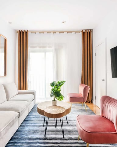 Modern living room with beige sofa, pink armchairs, wooden mirrors, wall-mounted TV, and glass doors with mustard curtains