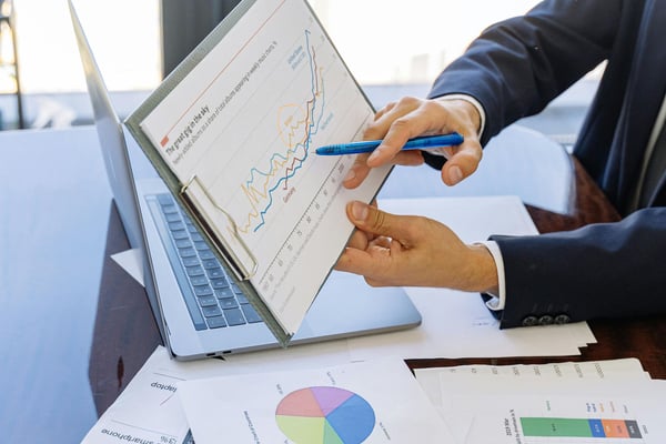 Business professionals reviewing financial charts and graphs on tablet and printed documents at desk with laptop and windows in background