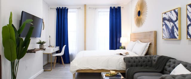 Modern bedroom with queen bed, gray tufted sofa, wall-mounted TV, blue curtains, and green plant
