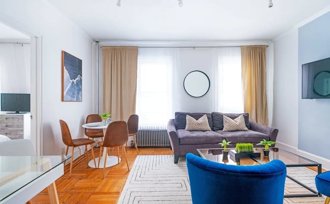 Bright living room with purple sofa, blue armchair, wooden dining table, hardwood floors, and large windows with tan curtains