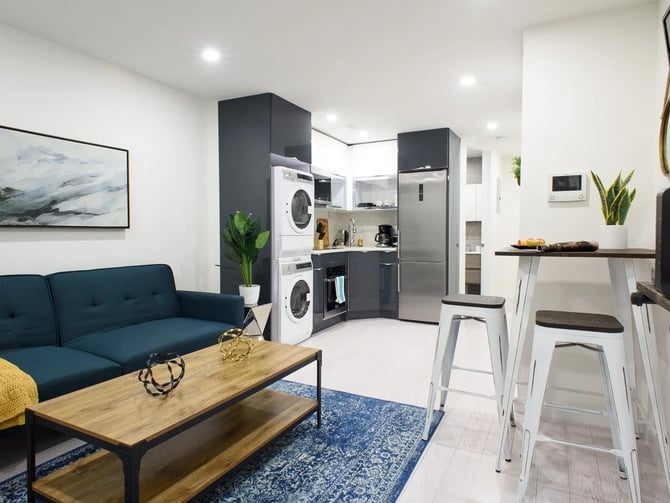 Open-plan studio with teal sofa, kitchen with stacked washers and dryers, bar seating, and stainless steel refrigerator.