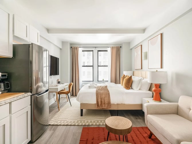 Modern studio apartment with white cabinetry, stainless steel appliances, queen bed with tan accents, and seating area with orange accents